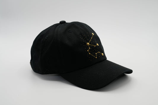 A black baseball cap with the Aquarius zodiac sign embroidered and riveted in gold on the front.