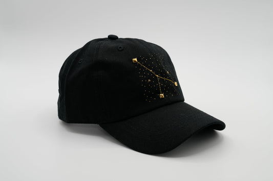 A black baseball cap with the Cancer zodiac sign embroidered and riveted in gold on the front.