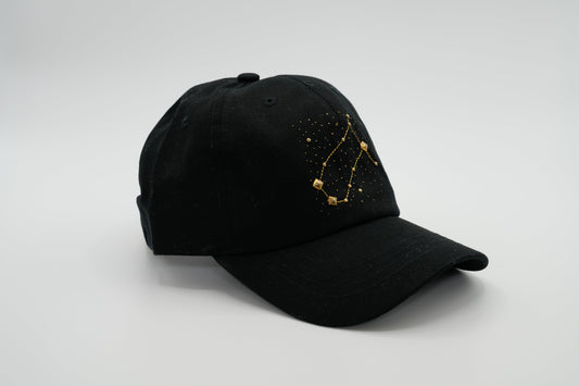 A black baseball cap with the Gemini zodiac sign embroidered and riveted in gold on the front.