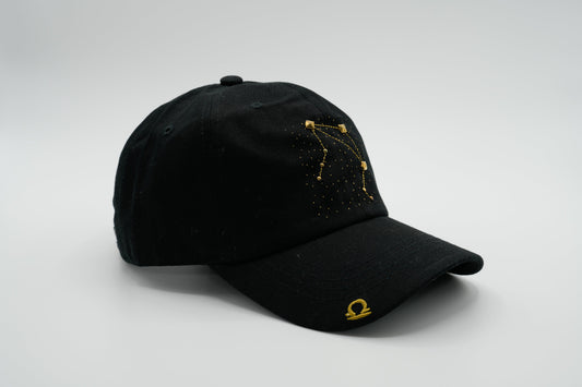 A black baseball cap with the Libra zodiac sign embroidered and riveted in gold on the front.