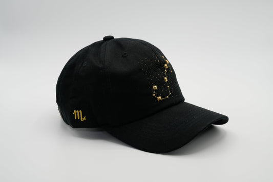 A black baseball cap with the Scorpio zodiac sign embroidered and riveted in gold on the front.