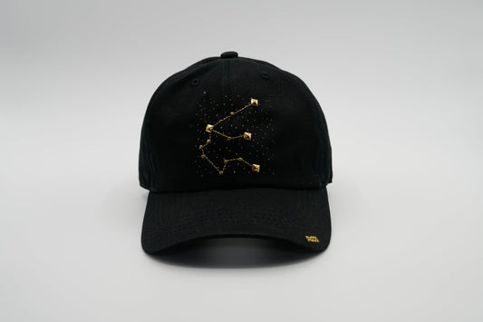 A black baseball cap with the Aquarius zodiac sign embroidered and riveted in gold on the front.