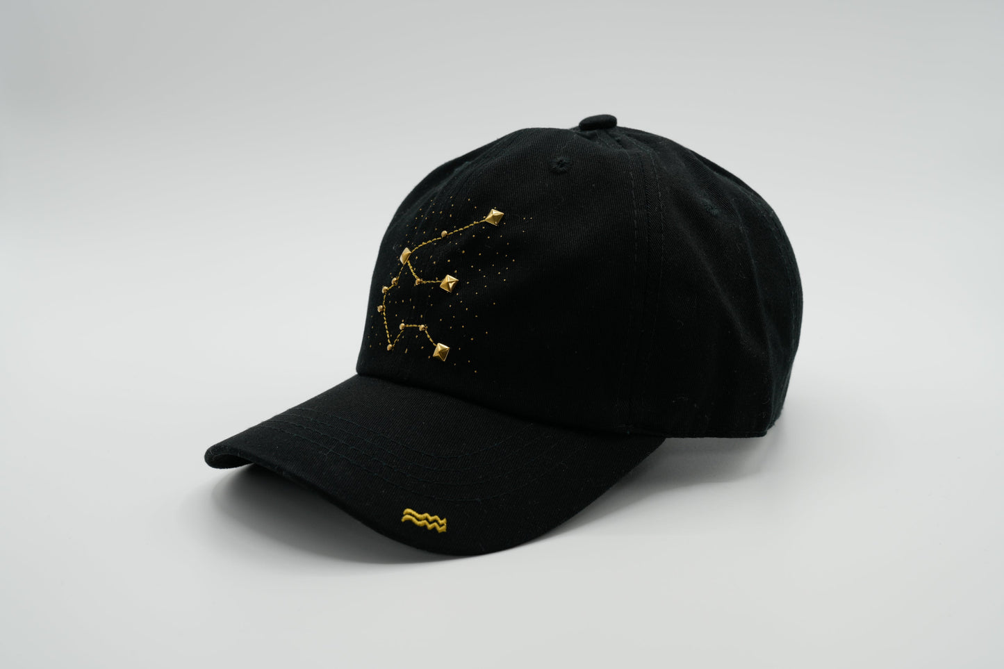 A black baseball cap with the Aquarius zodiac sign embroidered and riveted in gold on the front.