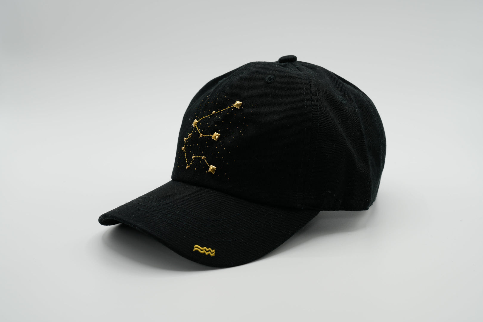 A black baseball cap with the Aquarius zodiac sign embroidered and riveted in gold on the front.