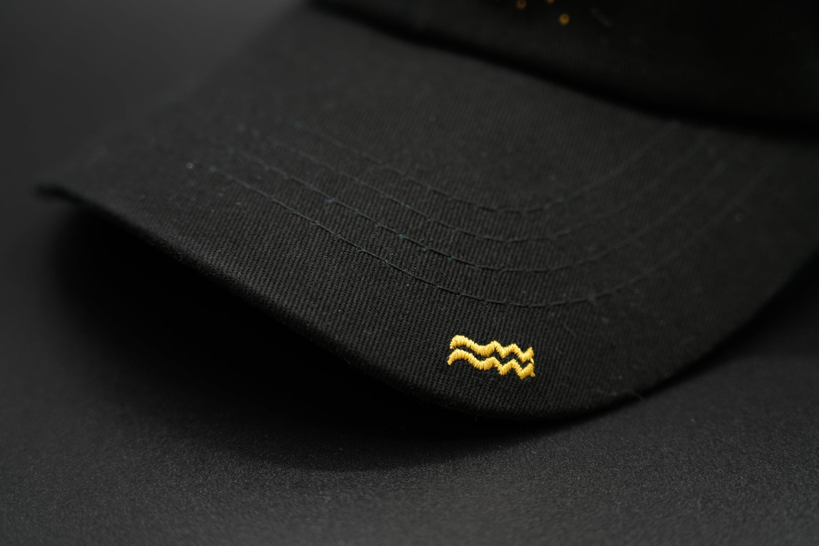 A black baseball cap with the Aquarius zodiac sign embroidered and riveted in gold on the front.