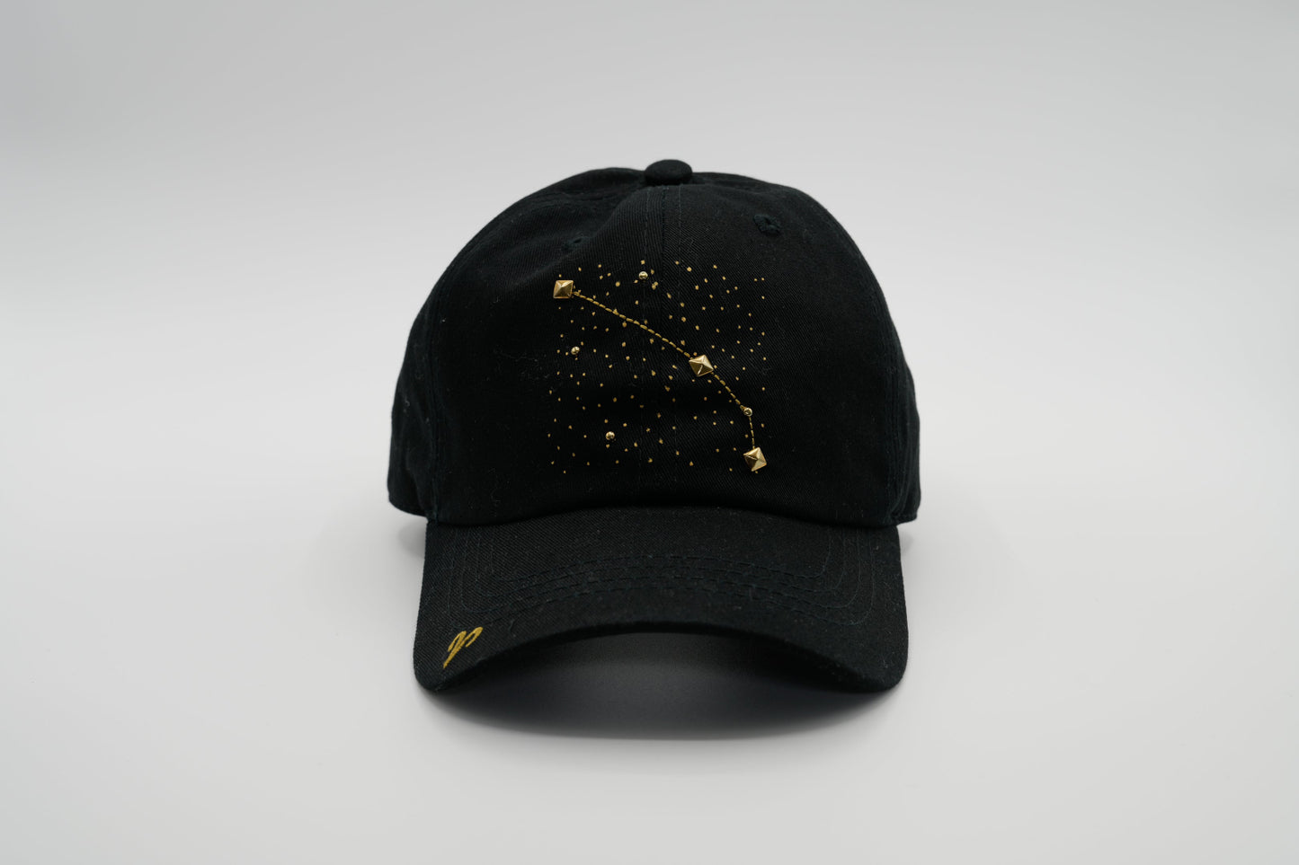 A black baseball cap with the Aris zodiac sign embroidered and riveted in gold on the front.