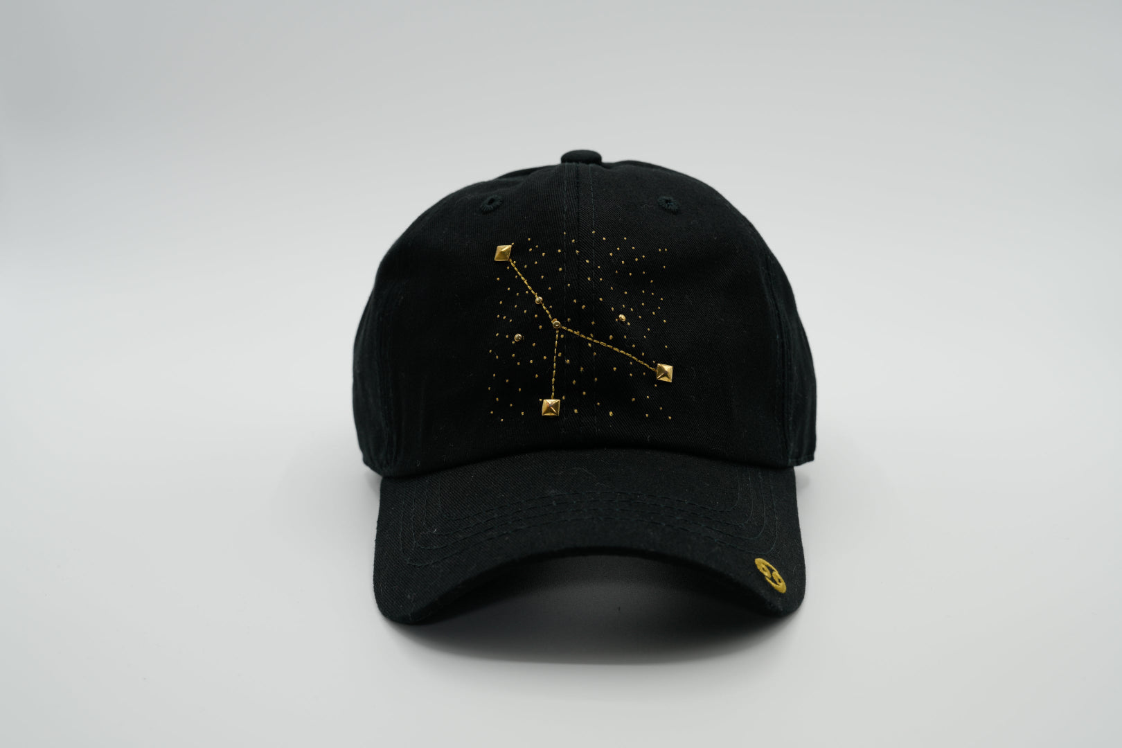 A black baseball cap with the Cancer zodiac sign embroidered and riveted in gold on the front.