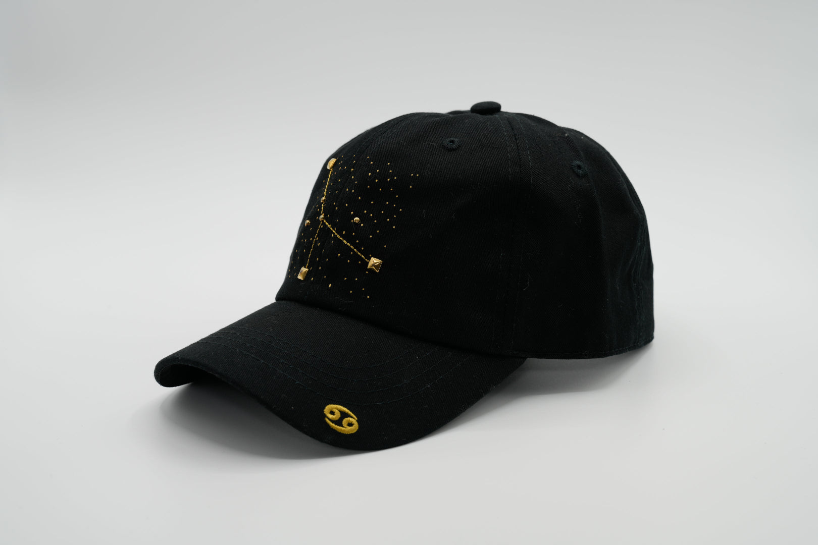 A black baseball cap with the Cancer zodiac sign embroidered and riveted in gold on the front.