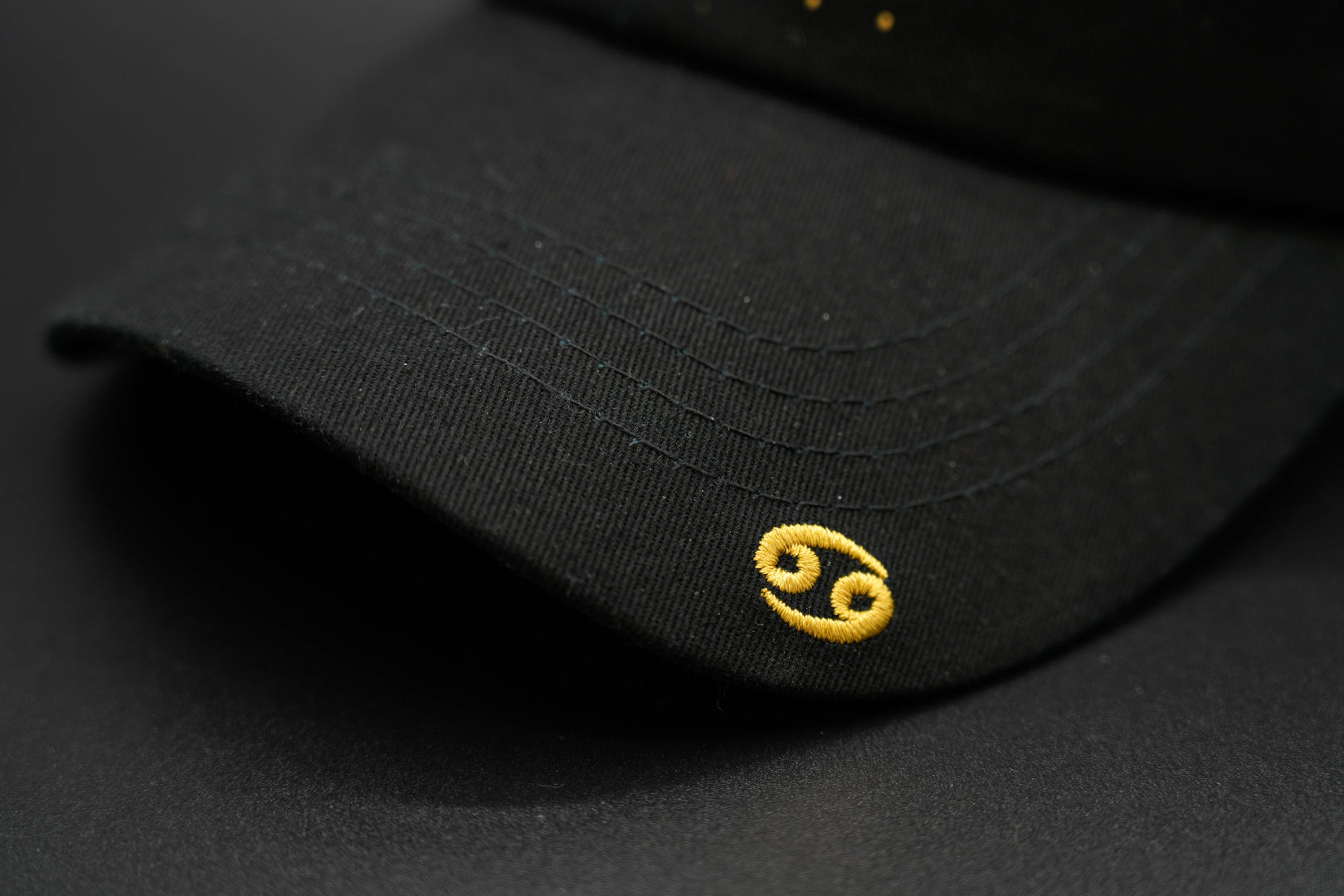 A black baseball cap with the Cancer zodiac sign embroidered and riveted in gold on the front.