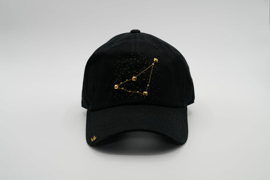 A black baseball cap with the Capricorn zodiac sign embroidered and riveted in gold on the front.