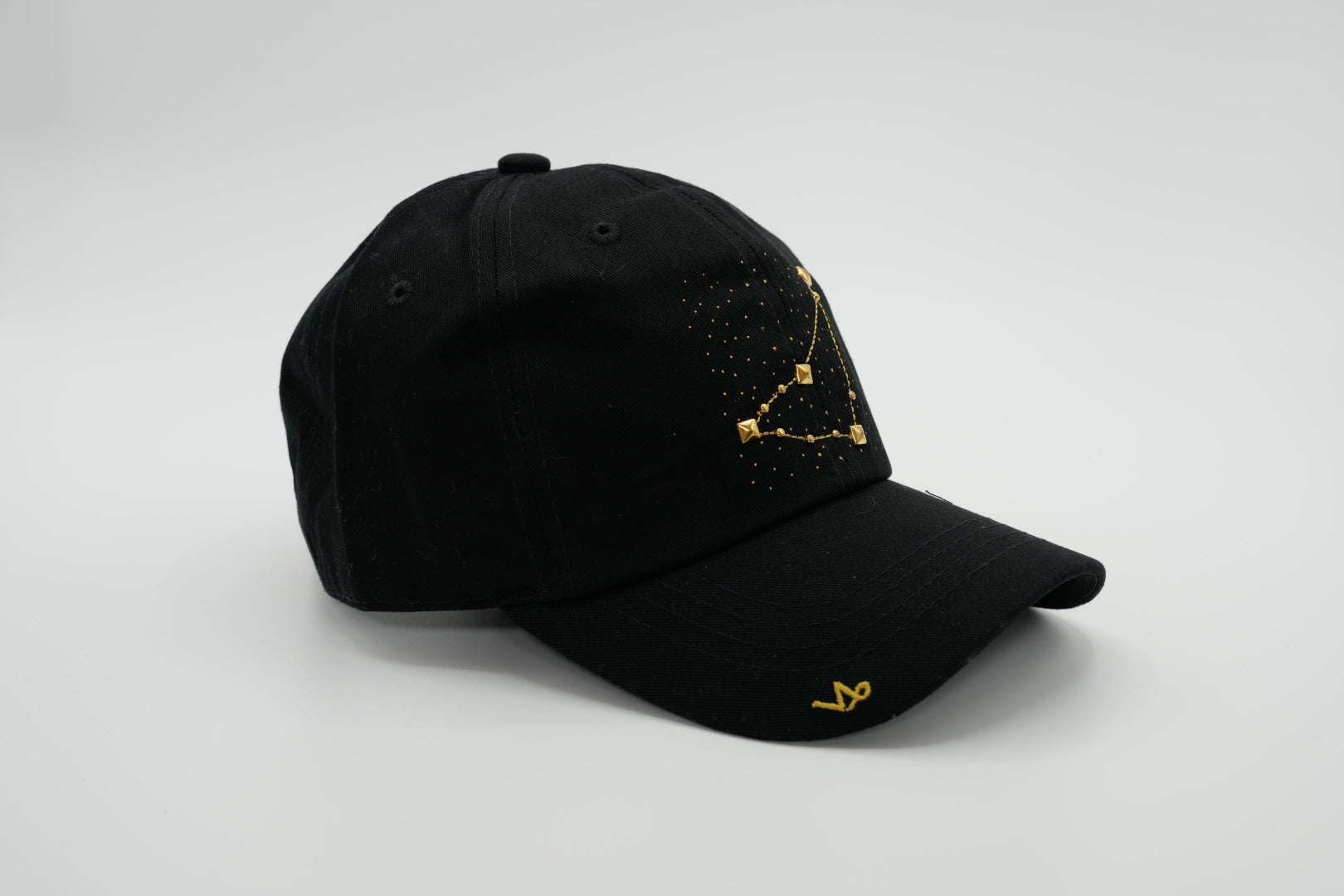 A black baseball cap with the Capricorn zodiac sign embroidered and riveted in gold on the front.