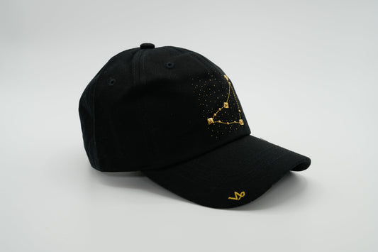 A black baseball cap with the Capricorn zodiac sign embroidered and riveted in gold on the front.