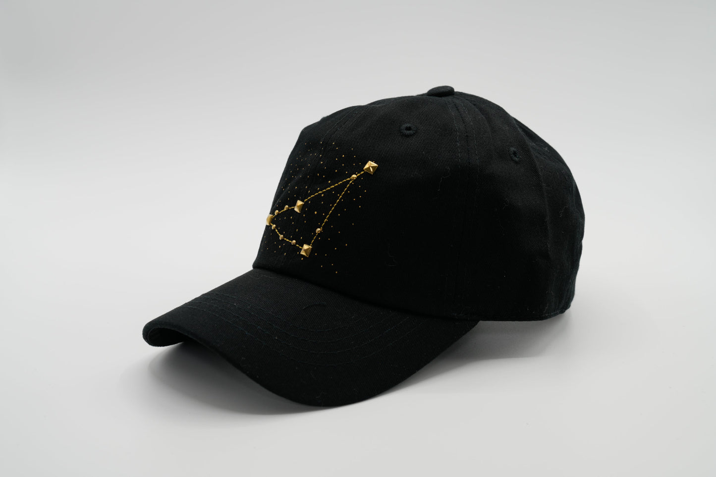 A black baseball cap with the Capricorn zodiac sign embroidered and riveted in gold on the front.