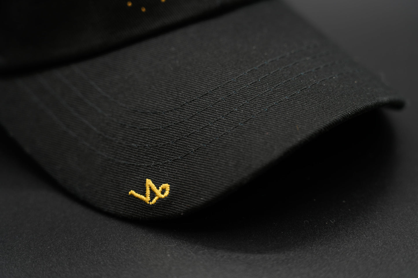 A black baseball cap with the Capricorn zodiac sign embroidered and riveted in gold on the front.