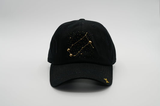A black baseball cap with the Gemini zodiac sign embroidered and riveted in gold on the front.