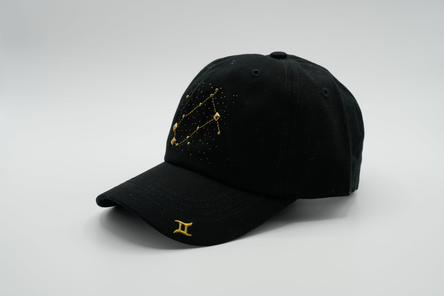 A black baseball cap with the Gemini zodiac sign embroidered and riveted in gold on the front.
