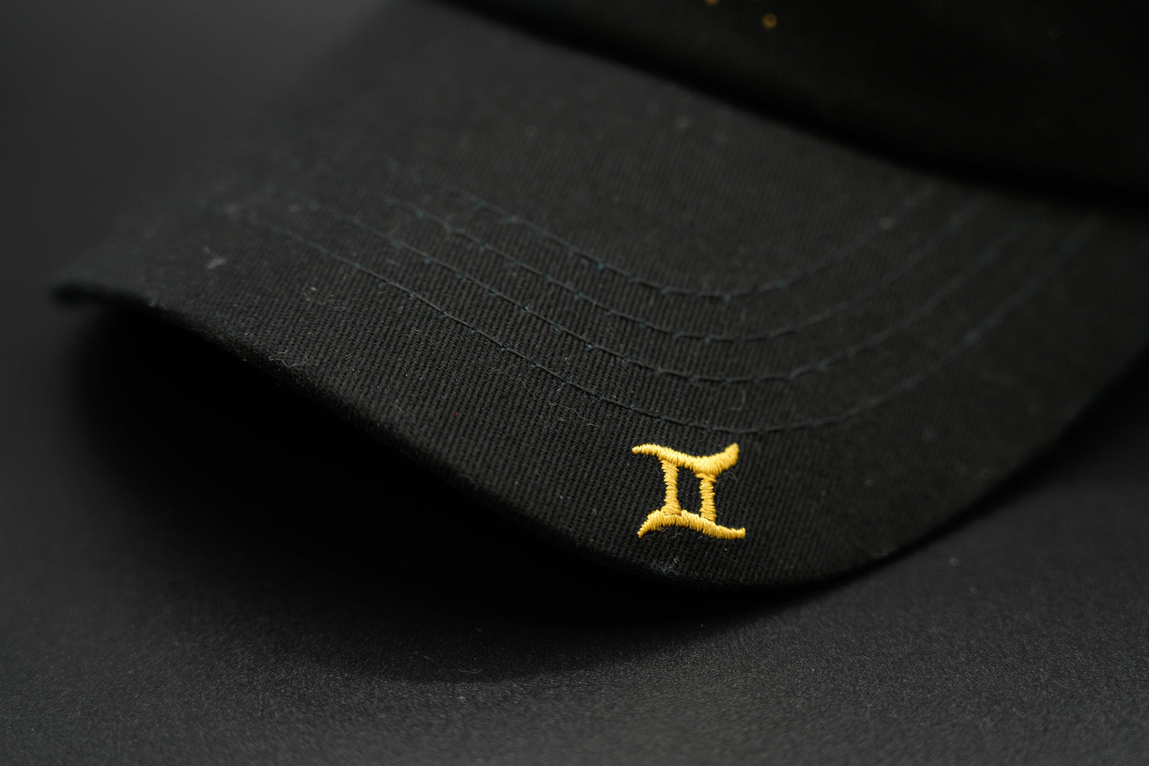 A black baseball cap with the Gemini zodiac sign embroidered and riveted in gold on the front.