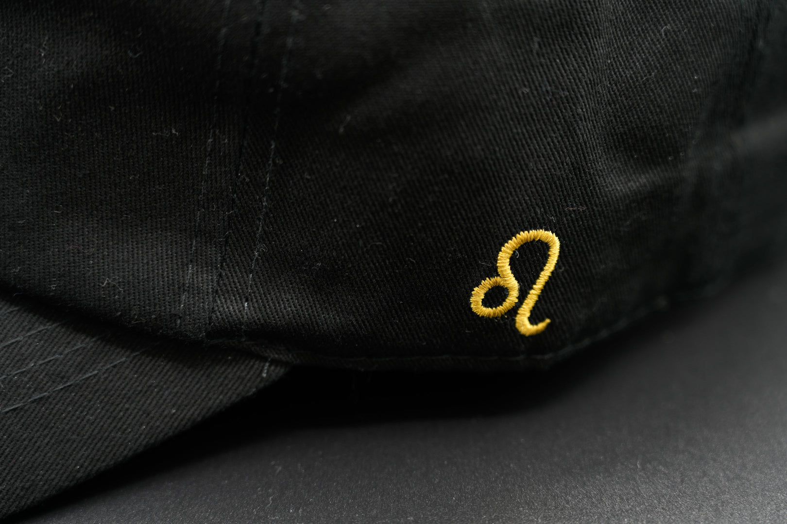A black baseball cap with the Leo zodiac sign embroidered and riveted in gold on the front.