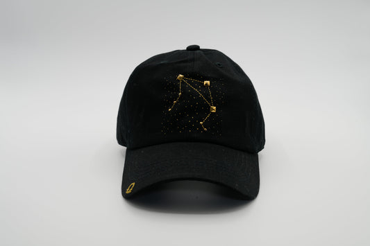 A black baseball cap with the Libra zodiac sign embroidered and riveted in gold on the front.