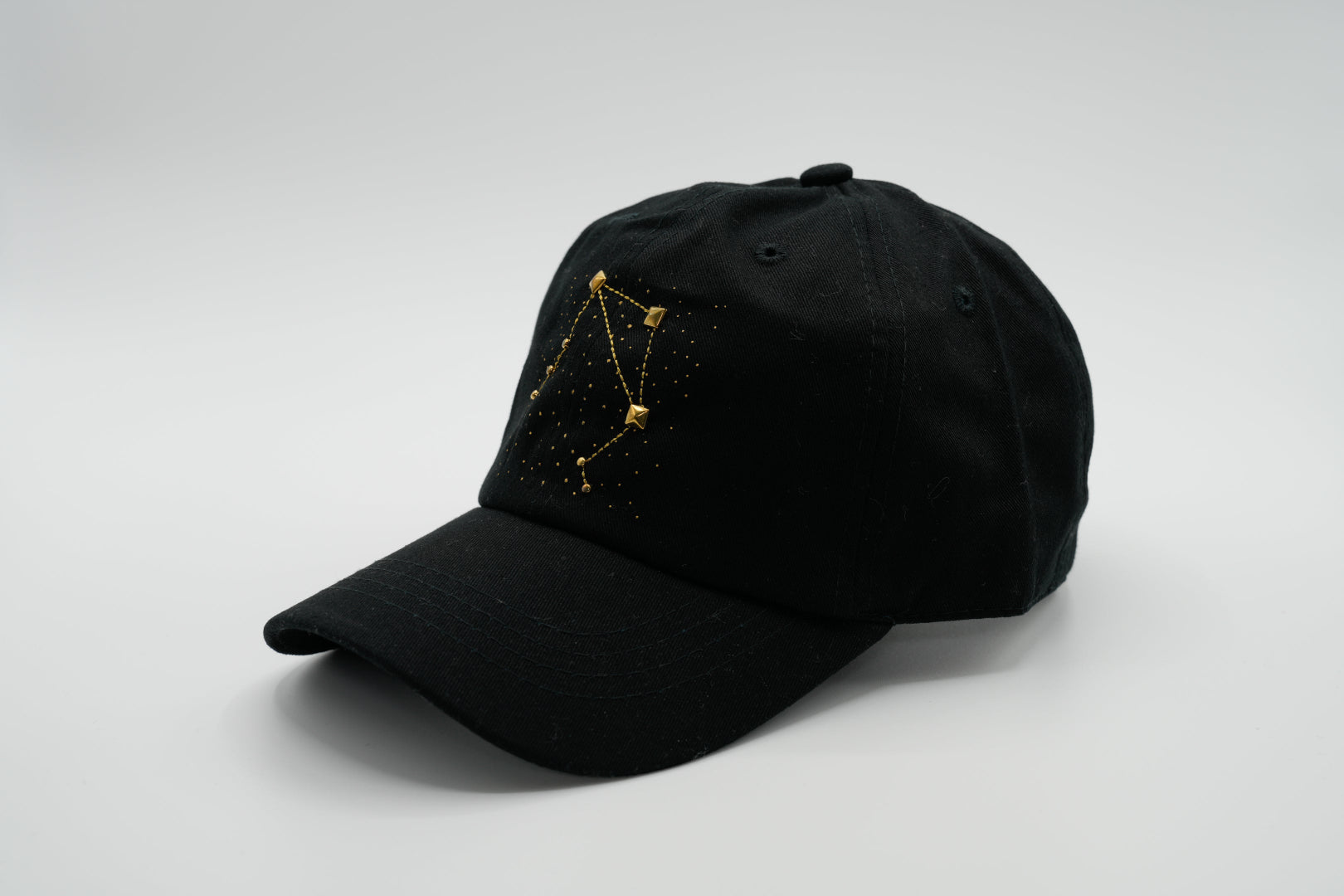 A black baseball cap with the Libra zodiac sign embroidered and riveted in gold on the front.