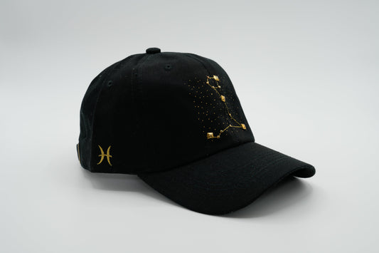 A black baseball cap with the Pisces zodiac sign embroidered and riveted in gold on the front.