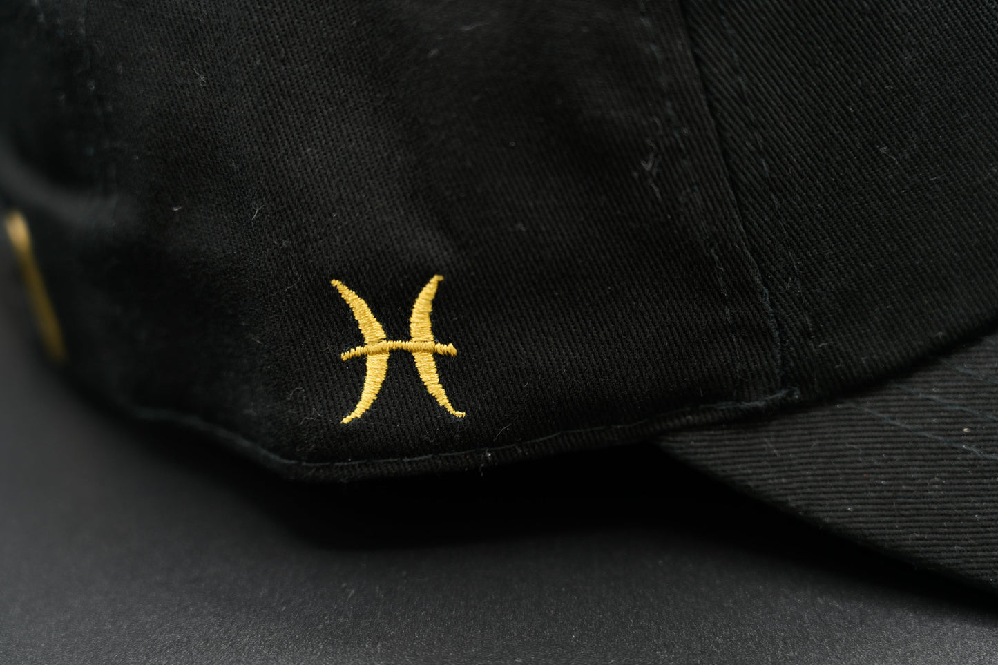 A black baseball cap with the Pisces zodiac sign embroidered and riveted in gold on the front.