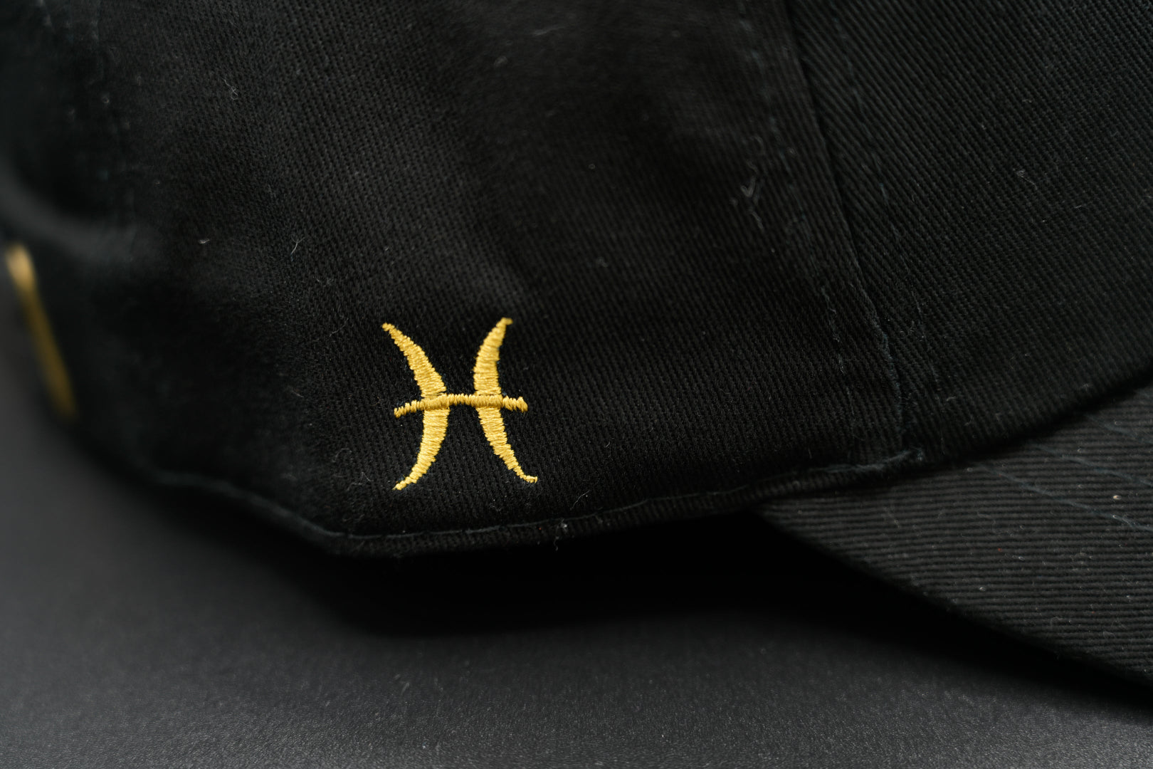 A black baseball cap with the Pisces zodiac sign embroidered and riveted in gold on the front.