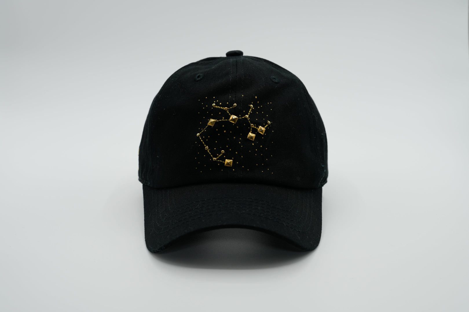 A black baseball cap with the Sagittarius zodiac sign embroidered and riveted in gold on the front.