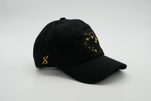 A black baseball cap with the Sagittarius zodiac sign embroidered and riveted in gold on the front.