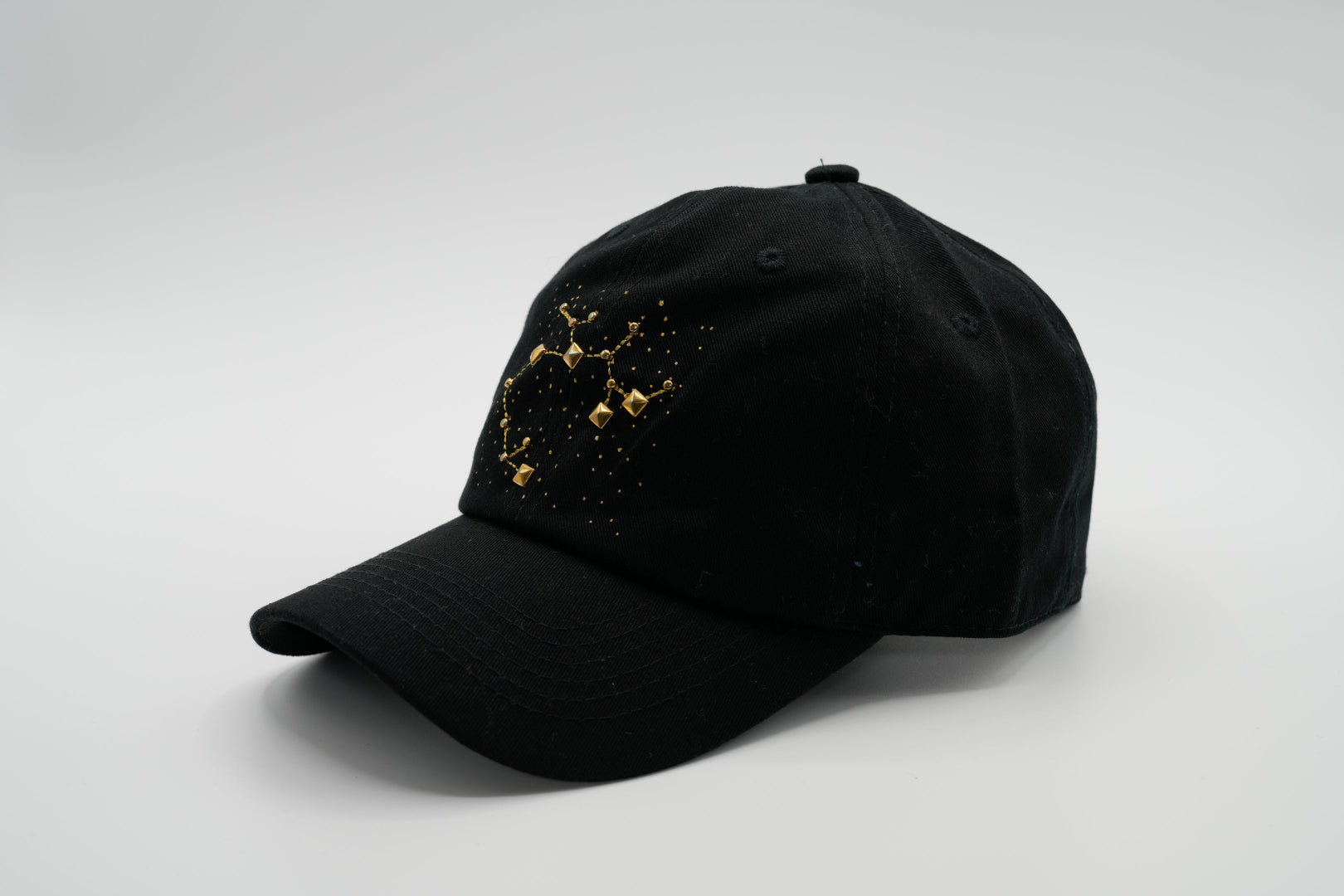 A black baseball cap with the Sagittarius zodiac sign embroidered and riveted in gold on the front.