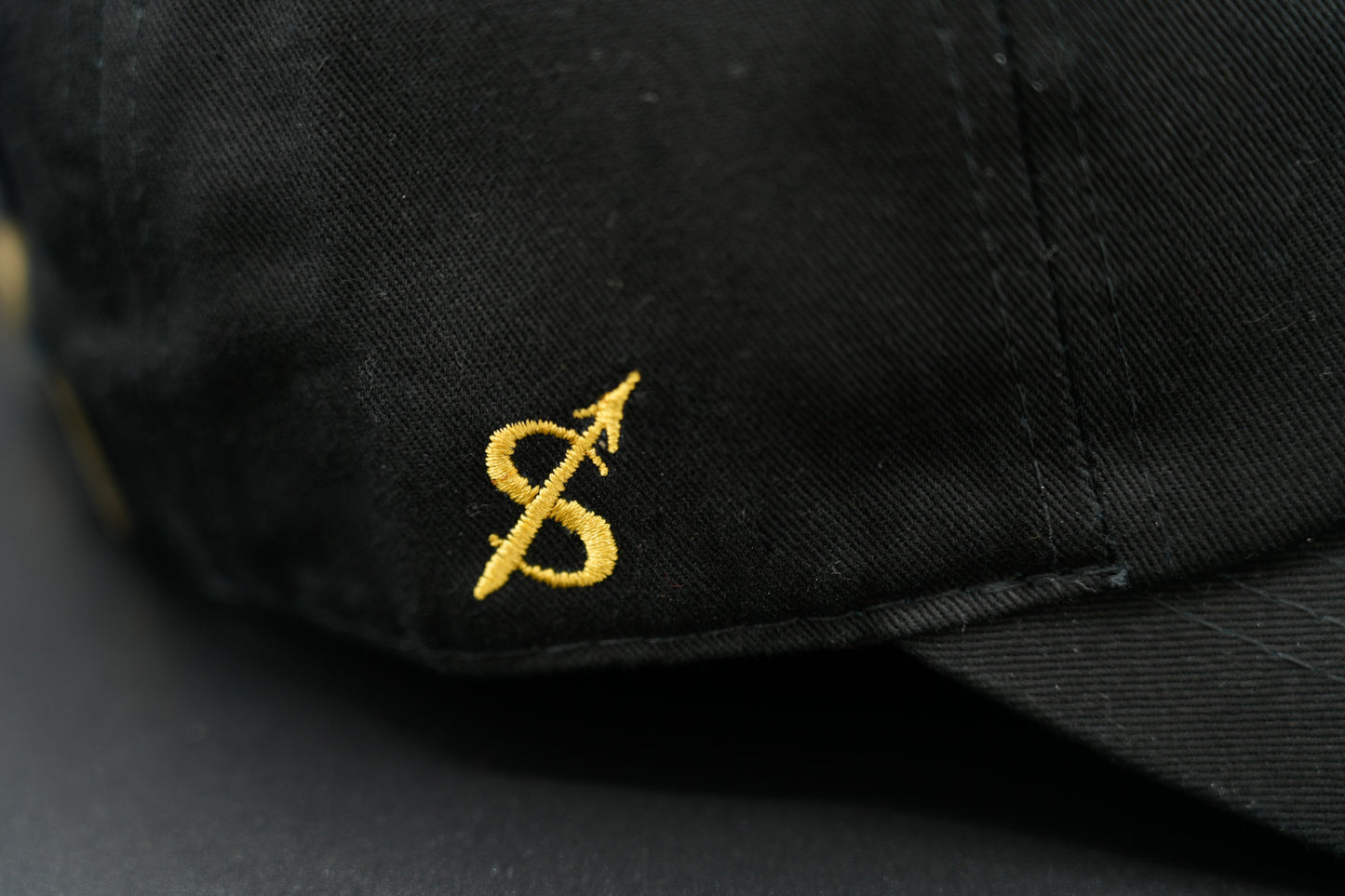 A black baseball cap with the Sagittarius zodiac sign embroidered and riveted in gold on the front.