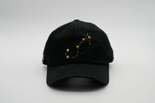 A black baseball cap with the Scorpio zodiac sign embroidered and riveted in gold on the front.