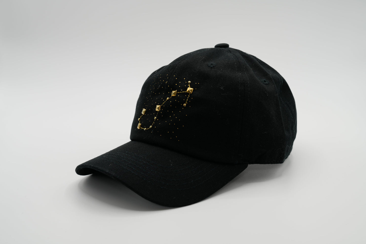 A black baseball cap with the Scorpio zodiac sign embroidered and riveted in gold on the front.