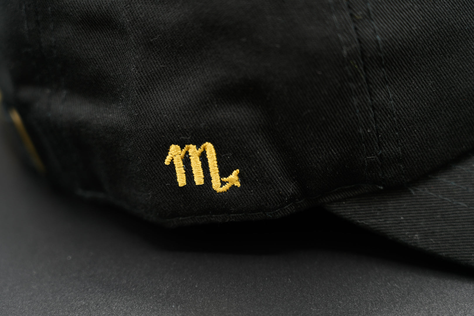A black baseball cap with the Scorpio zodiac sign embroidered and riveted in gold on the front.