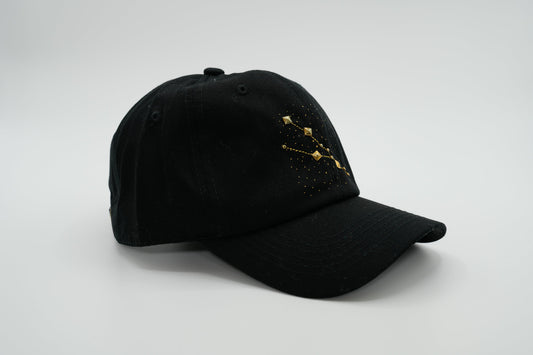 A black baseball cap with the Taurus zodiac sign embroidered and riveted in gold on the front.