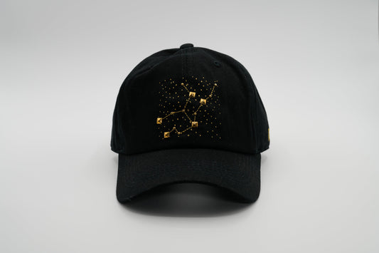 A black baseball cap with the Virgo zodiac sign embroidered and riveted in gold on the front.