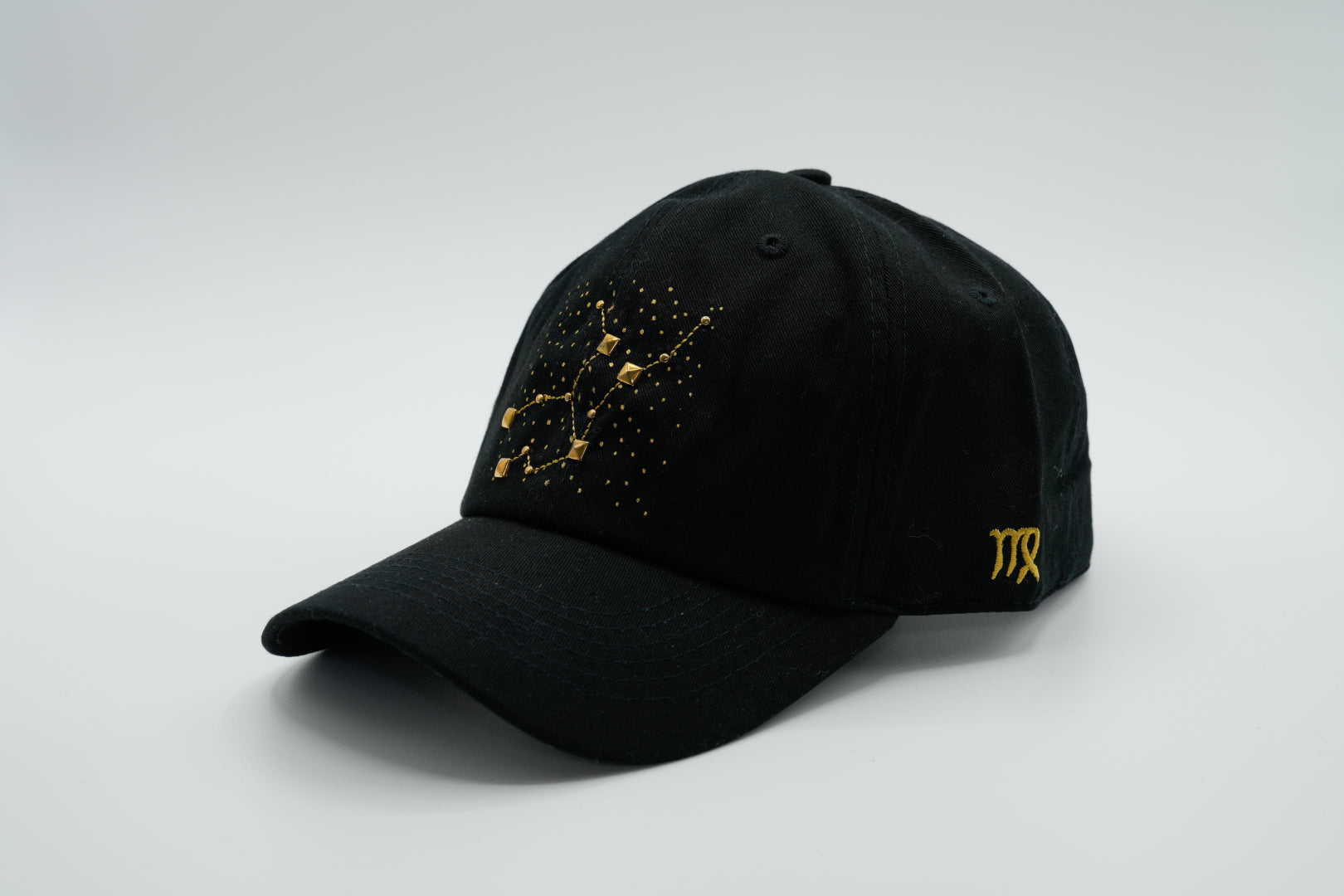 A black baseball cap with the Virgo zodiac sign embroidered and riveted in gold on the front.