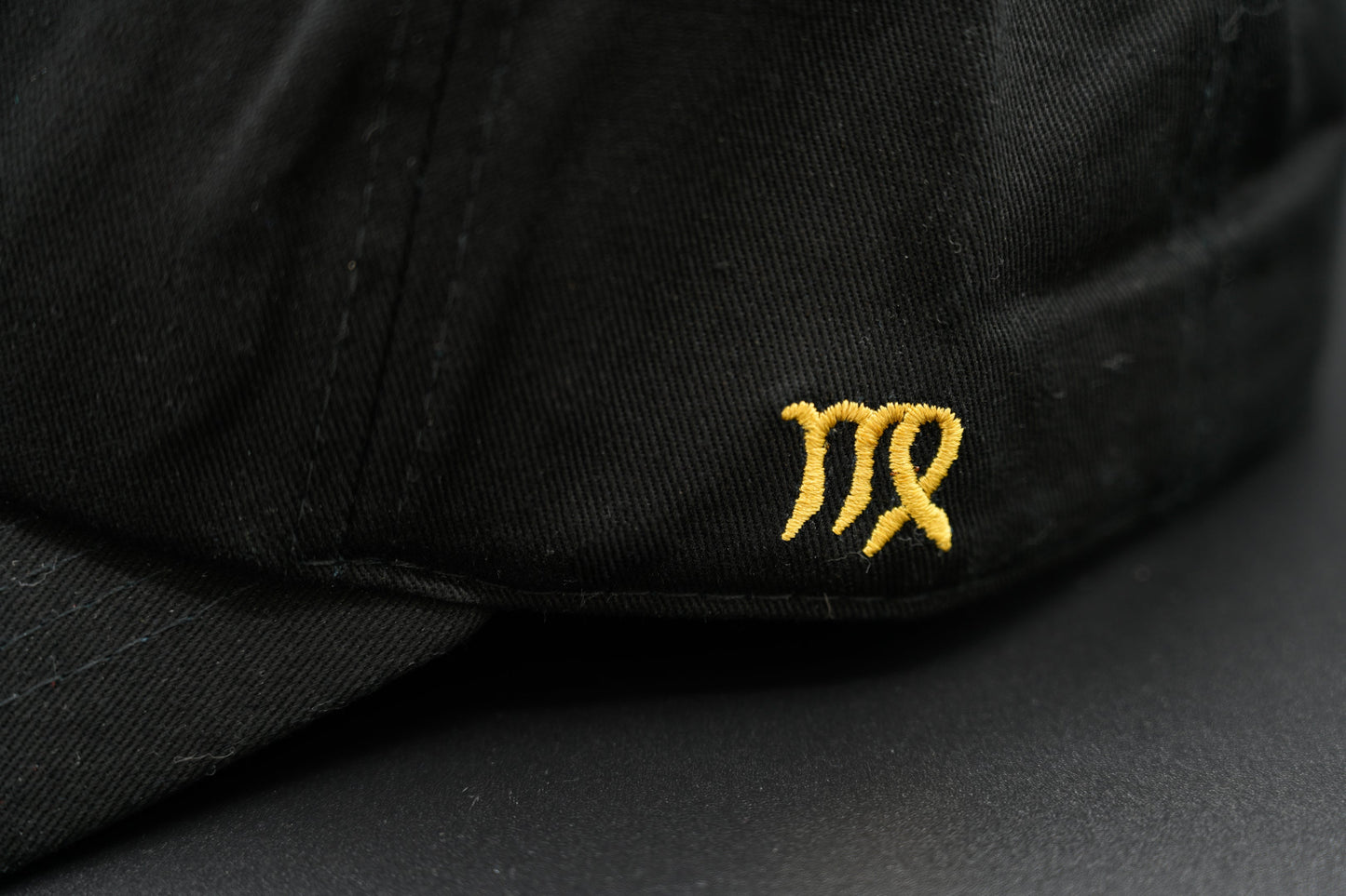A black baseball cap with the Virgo zodiac sign embroidered and riveted in gold on the front.