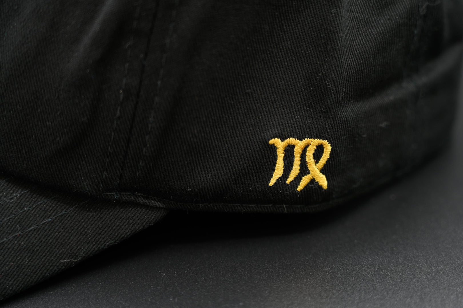 A black baseball cap with the Virgo zodiac sign embroidered and riveted in gold on the front.